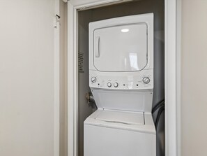 A white, compact washer and dryer unit with control knobs and a start button, installed in a corner with a door. - Edgewater Leeward #302 | Gulf Front (Panama City Beach)
