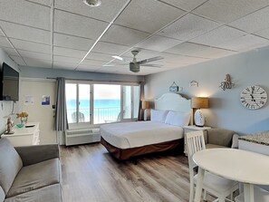 Ein Hotelzimmer mit Bett, Schreibtisch, Stuhl, Deckenventilator und Blick auf das Meer durch die Fenster.