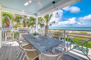 Un patio techado al aire libre con una parrilla, una mesa de comedor y sillas, con vista a una playa con palmeras y el océano.