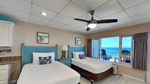 Ein Hotelzimmer mit zwei Betten, einem Deckenventilator, einem Fenster mit Blick auf den Ozean und einem Wandbild im Strandhausstil.