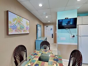 A dining area with a table set for four, a television mounted on the wall, and a refrigerator to the right.