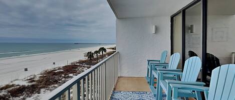 A balcony with a view of the beach and a pier, featuring light blue Adirondack chairs and a patterned rug.