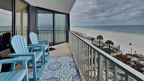 A balcony with two blue Adirondack chairs, a patterned rug, and a view of the beach and ocean. - Edgewater Leeward #306 | Gulf Front + Amenities (Panama City Beach)