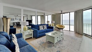 A spacious living room with a blue sofa set, a white coffee table, and a yellow chair. There is a large window offering a view of the ocean. - Edgewater Leeward #306 | Gulf Front + Amenities (Panama City Beach)
