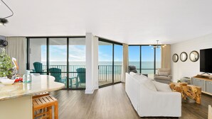 Uma sala de estar moderna com um sofá branco, uma mesa de centro de madeira e uma televisão. Há uma varanda com vista para o oceano.