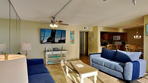 A living room with a ceiling fan, a TV, a coffee table, and a sofa. There is a kitchen in the background. - Edgewater Leeward #407 | Gulf Front + Amenities (Panama City Beach)