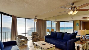 A spacious living room with a large window overlooking the sea, a ceiling fan, and a blue sofa. - Edgewater Leeward #407 | Gulf Front + Amenities (Panama City Beach)