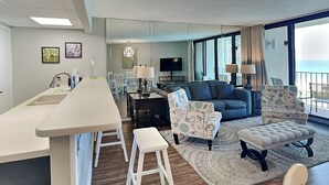 Una sala de estar moderna con un sofá, dos sillones estampados, una mesa de centro y un comedor con una mesa larga y sillas.