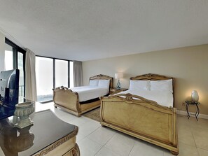 A hotel room with two ornate beds, a small table, a lamp, and a view of the city through large windows.