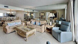 Amplia sala de estar con un conjunto de sofá color beige, una mesa de centro de vidrio y un sillón azul. Un espejo grande refleja la habitación, y se distingue un comedor al fondo.