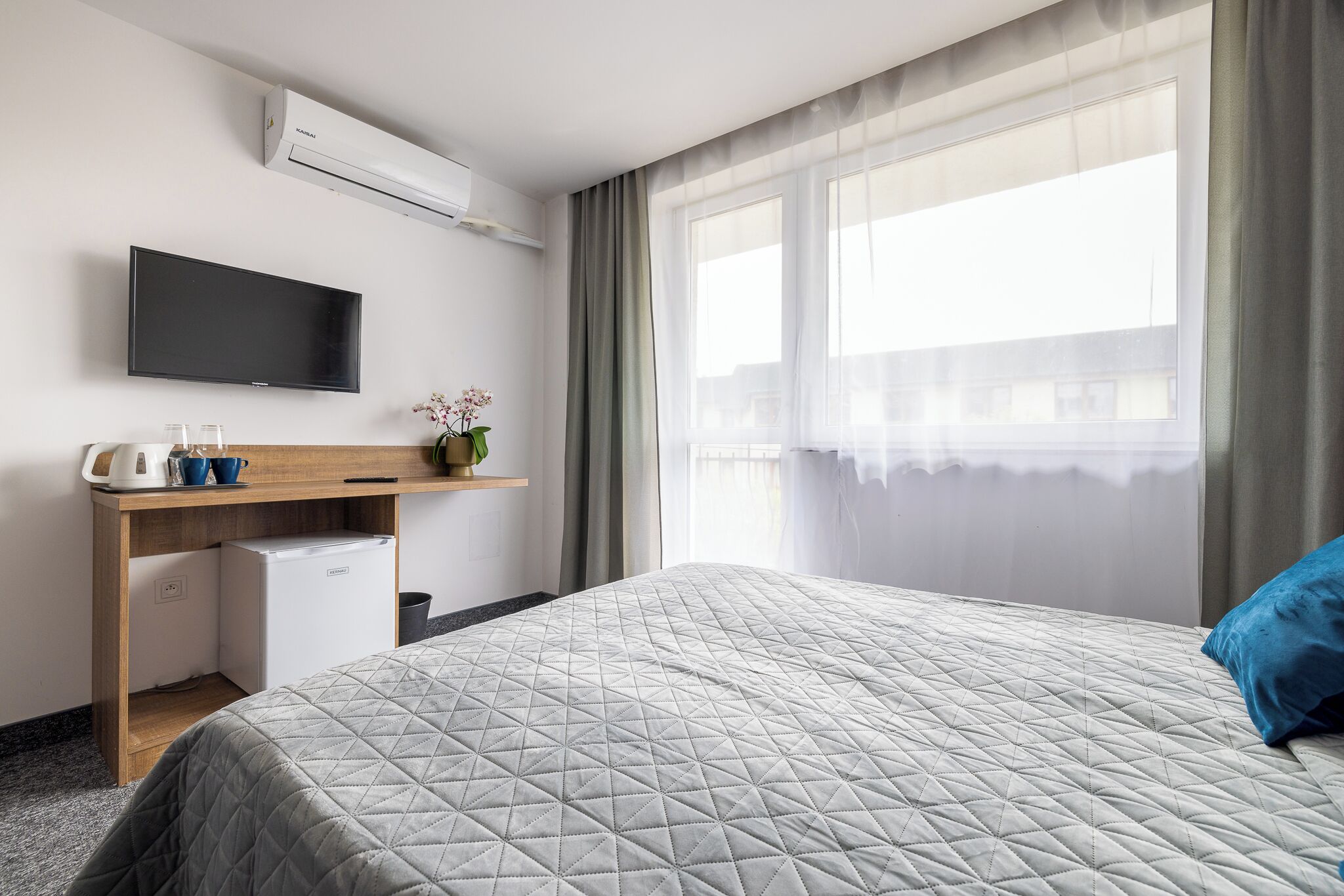 Een hotelkamer met een bed, een aan de muur bevestigde televisie, een klein bureau met een koelkast en een raam met gordijnen.
