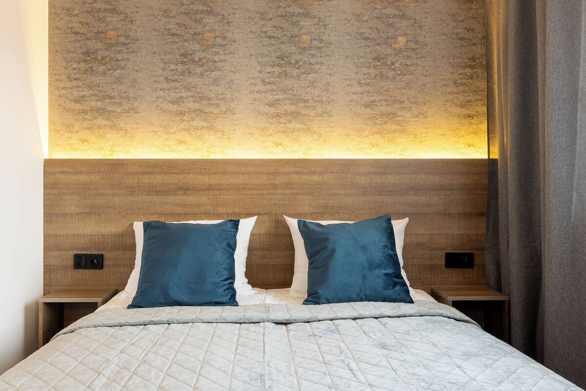 Ein ordentlich bezogenes Bett mit einer gesteppten Decke, zwei Kissen, einem Holzkopfstück und einer Wand mit strukturierter Oberfläche.
