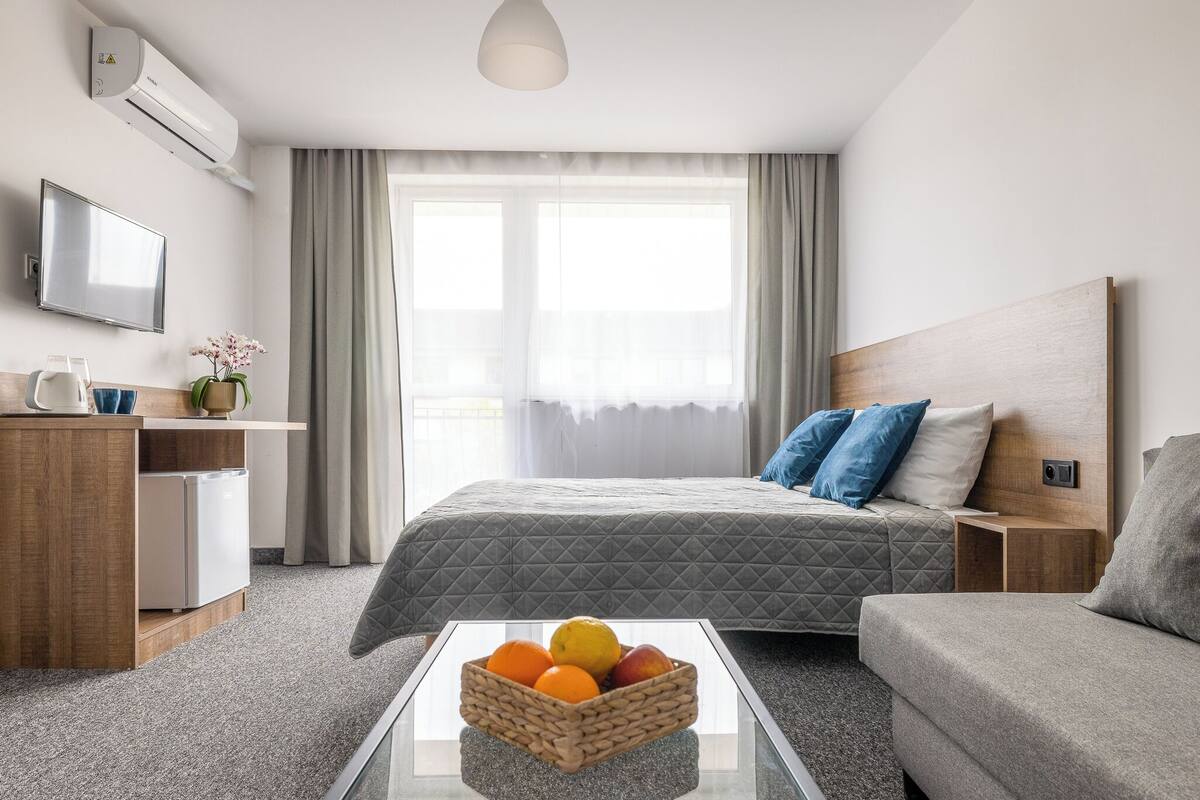 Ein modernes Hotelzimmer mit einem Bett, einer Couch, einem Couchtisch mit Obst, einem Fernseher, einer Minibar und einem Fenster mit Vorhängen.