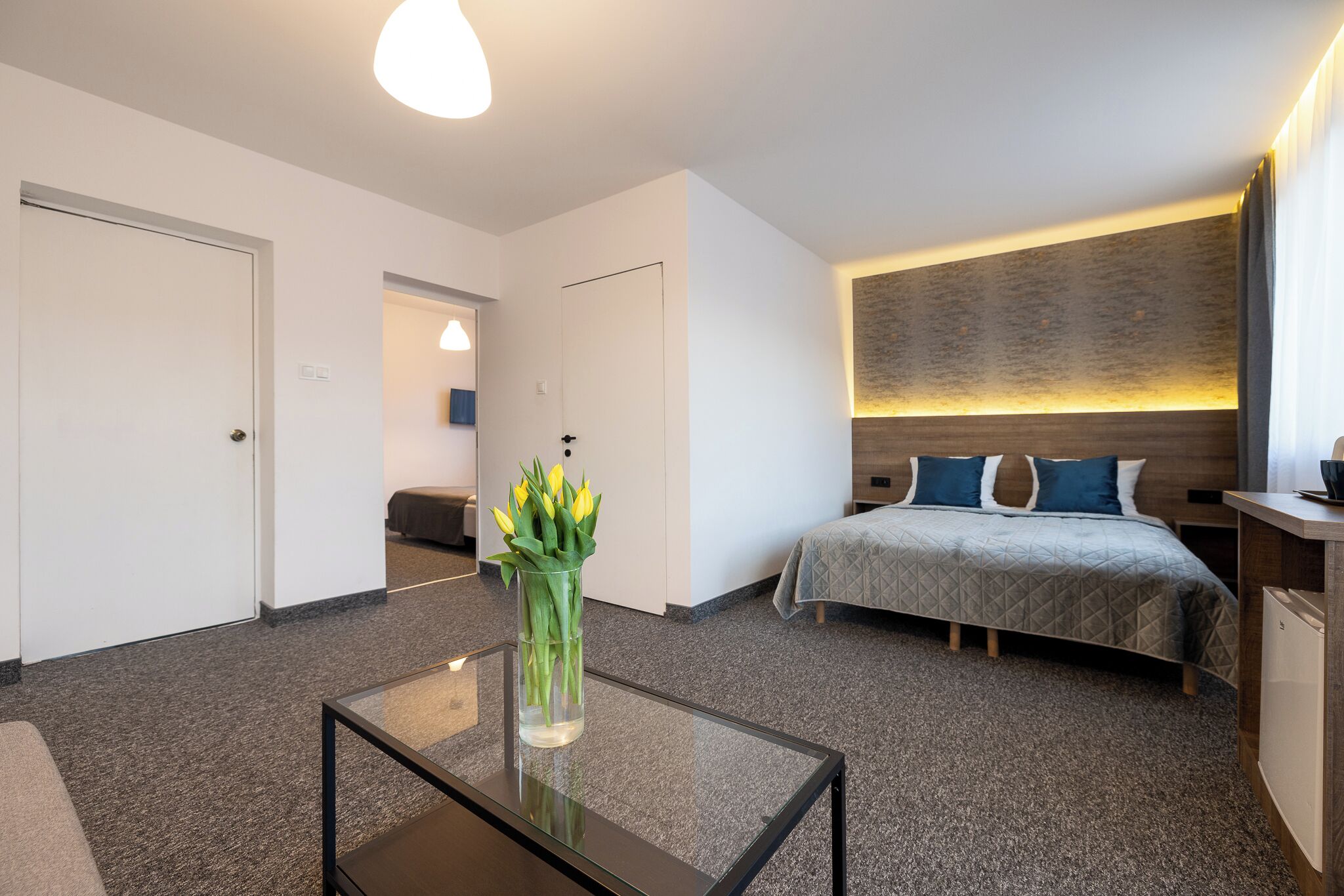 Une chambre d’hôtel moderne équipée d’un lit, d’une table de chevet et d’une table basse en verre sur laquelle est posé un vase de fleurs.