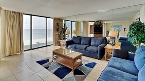 Un salon spacieux doté d’une grande fenêtre donnant sur la plage, d’un canapé bleu, d’une table basse et d’un téléviseur.