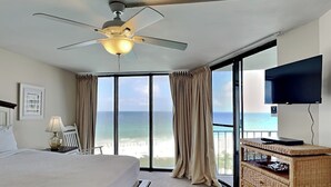Habitación de hotel con una cama, ventilador de techo, televisión, cómoda de mimbre y un balcón con vista al océano.
