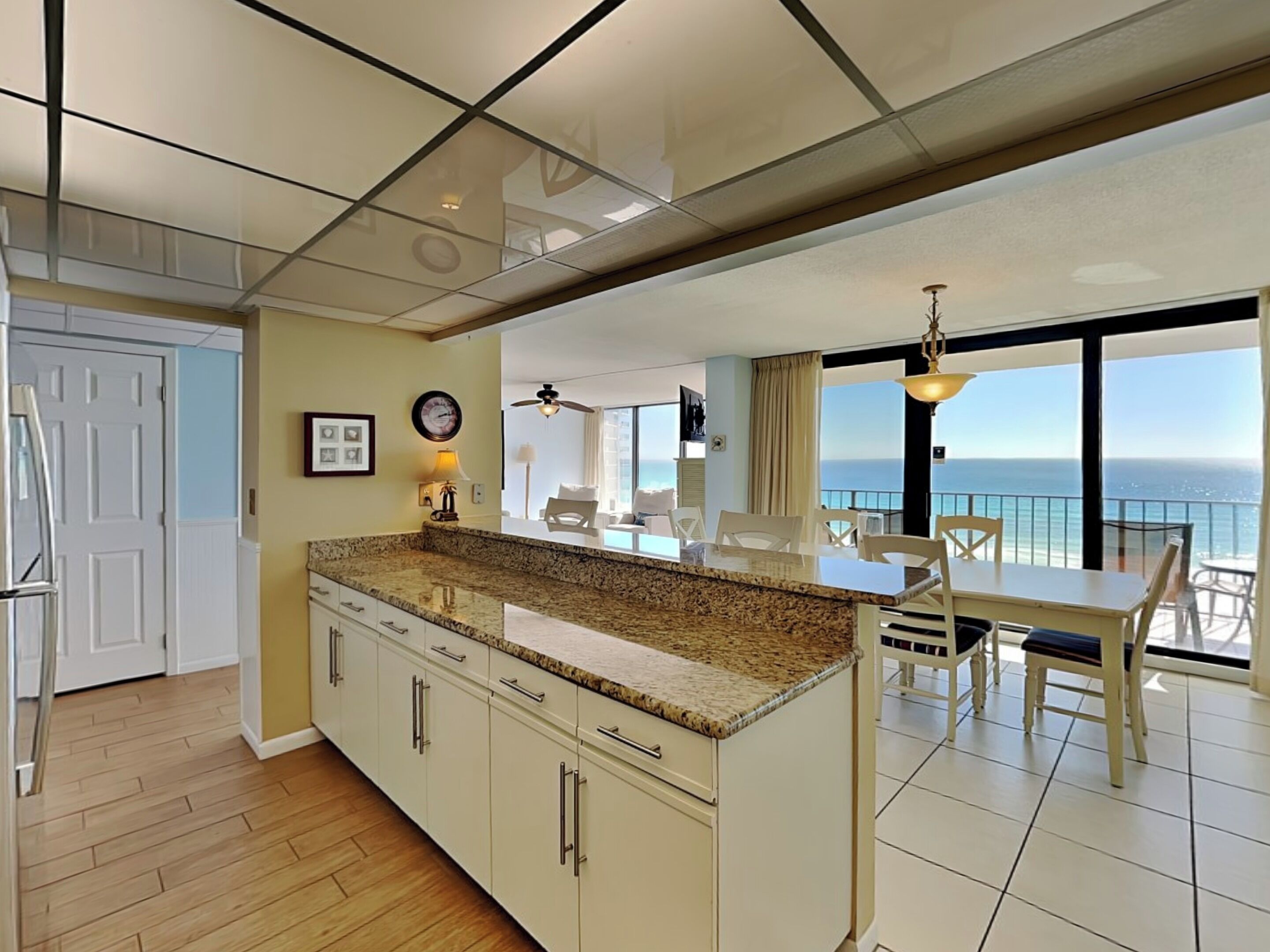 A modern kitchen with granite countertops, white cabinets, and a large island. There is a dining area with a table and chairs, and a balcony with a view of the ocean.