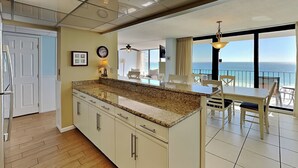 A modern kitchen with granite countertops, white cabinets, and a large island. There is a dining area with a table and chairs, and a balcony with a view of the ocean.