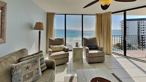A spacious living room with a view of the beach, a ceiling fan, and a large window.