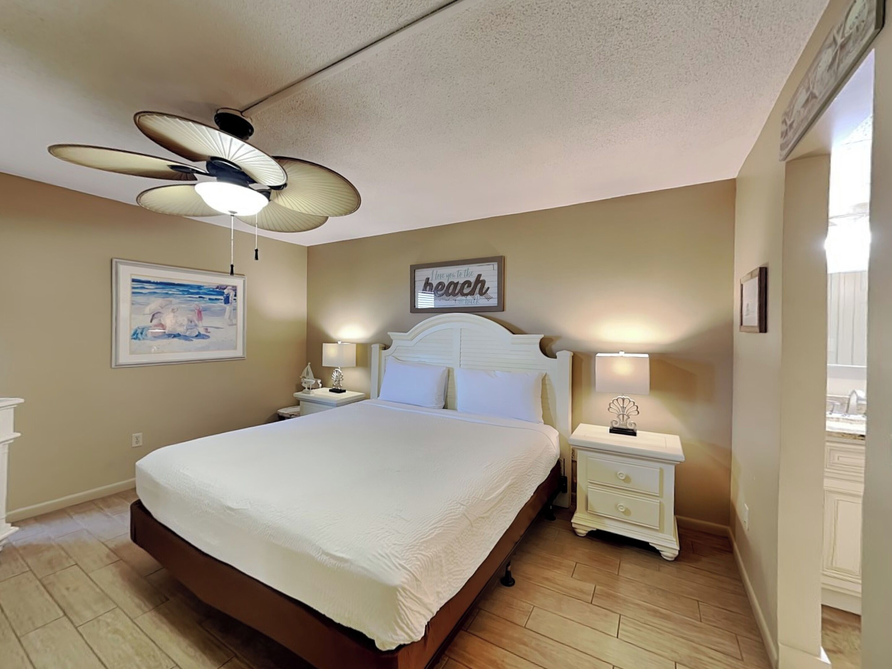 Une chambre à coucher comprenant un lit, deux tables de chevet, un ventilateur de plafond et un mur orné d’un tableau encadré et d’une affiche.