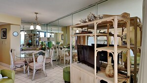 A spacious living room with a large glass dining table, wicker chairs, a green ottoman, and a wooden entertainment unit with shelves.