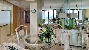 A dining area with wicker chairs, a glass table, and a vase of flowers. There is a clock on the wall and a view of the sea through large windows.