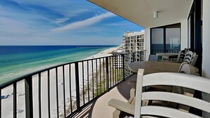 A balcony with a view of the beach and ocean, a table with chairs, and a building in the background. - Edgewater Tower II #1001 | Amenities & Views Galore! (Panama City Beach)