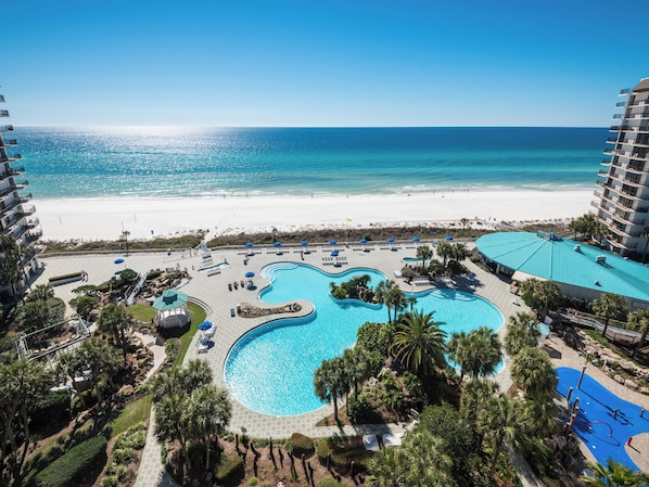 Relax by the winding pool surrounded by lush greenery and palm trees, with a scenic view of the ocean and sandy beach.
