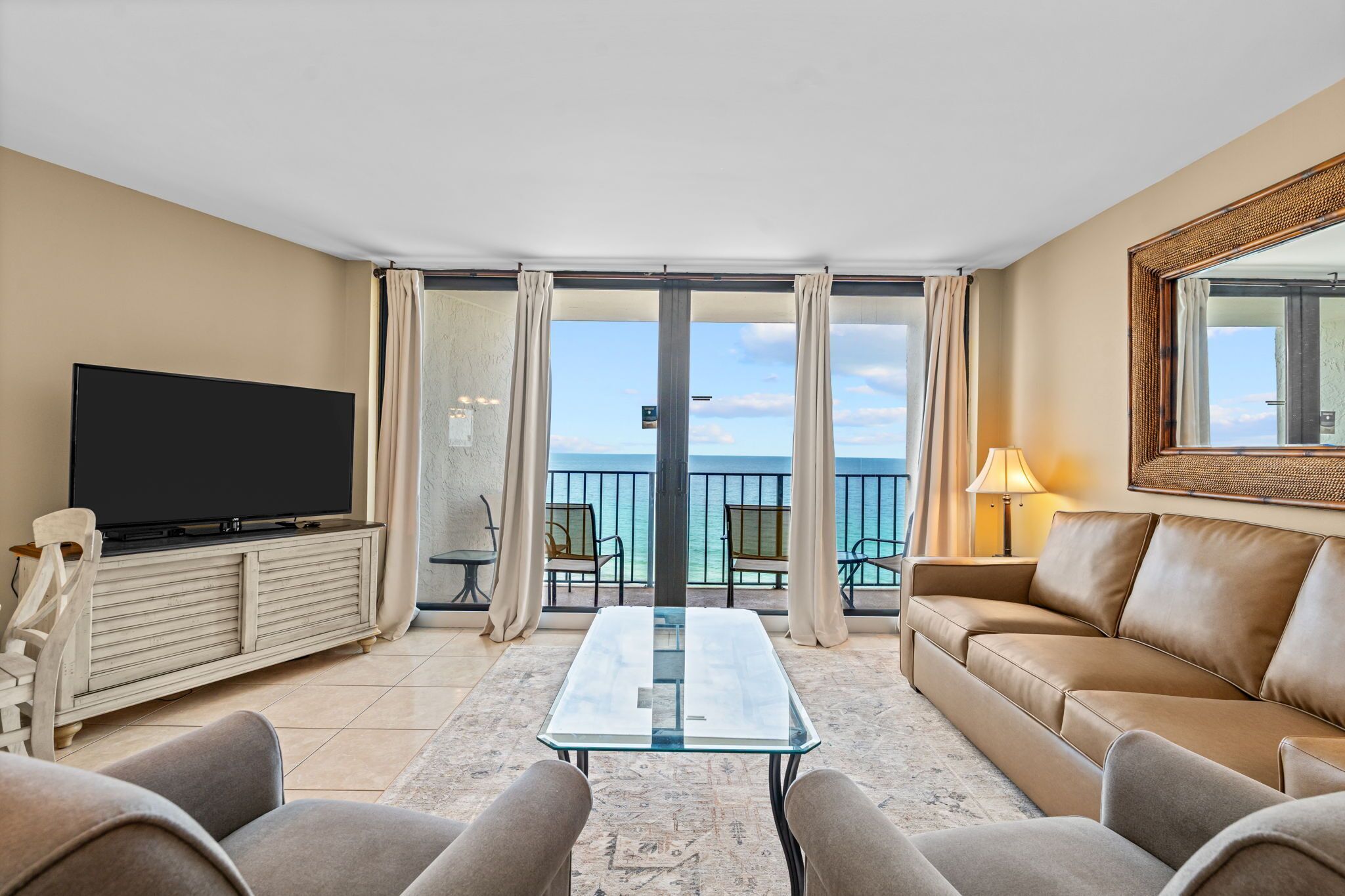 Habitación de hotel con televisor, sofá, mesa de vidrio y balcón con vista al mar.