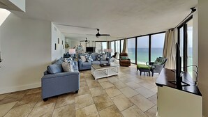 Un vaste espace de vie comprenant un canapé, une table basse et des fauteuils, de grandes fenêtres offrant une vue sur l’océan et un ventilateur de plafond.