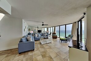 A spacious living area with a sofa, coffee table, and chairs, large windows with a view of the ocean, and a ceiling fan.
