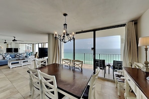 Een ruime woonkamer met een eettafel, witte stoelen, een kroonluchter en een balkon met uitzicht op zee.