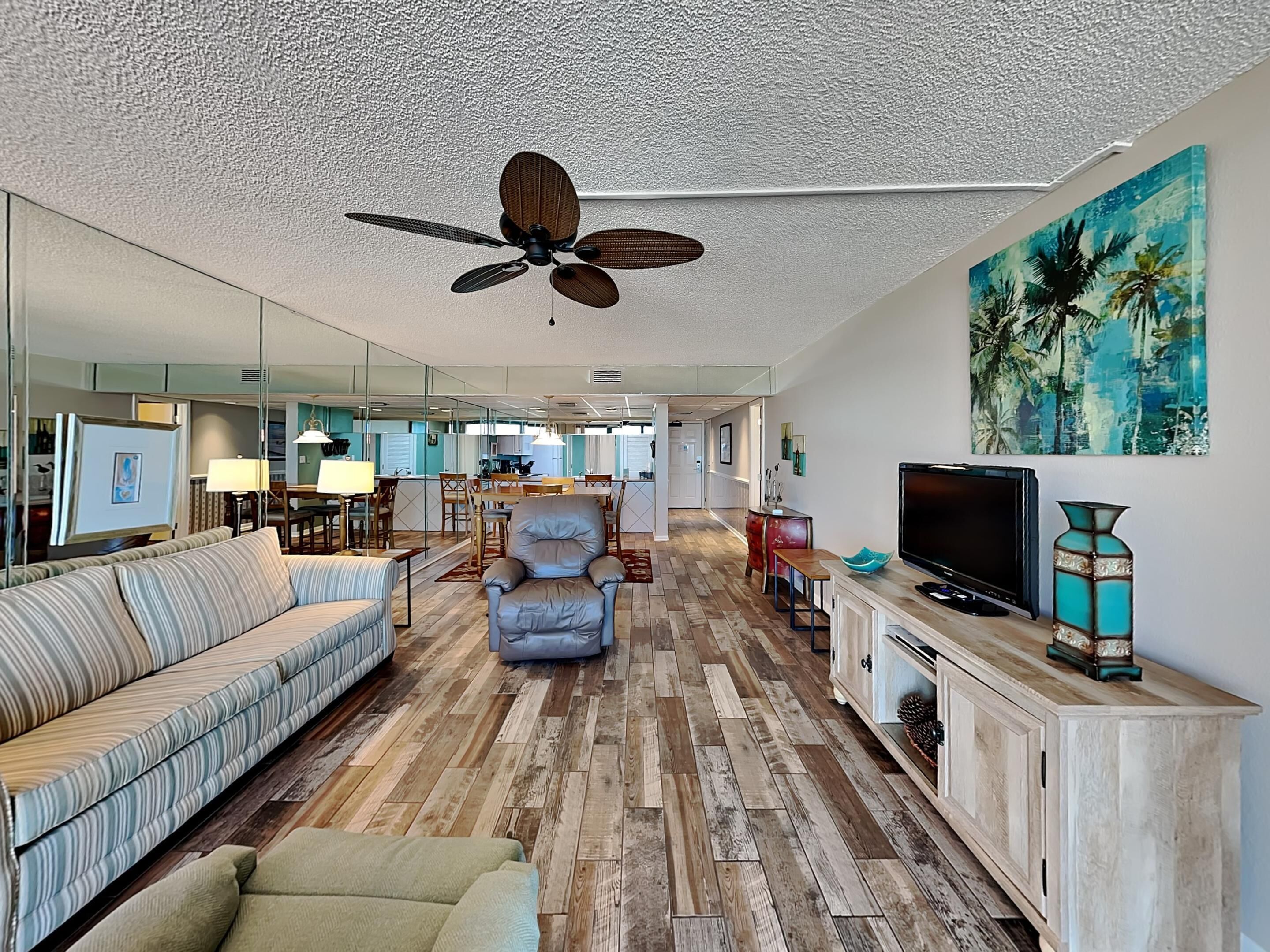 A spacious living room with a ceiling fan, a TV, and a painting of palm trees.