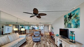 Un salón amplio con ventilador de techo, televisor y un cuadro de palmeras.