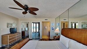 Habitación de hotel con una cama grande, un ventilador de techo, un televisor sobre una cómoda y vistas a la playa a través de las puertas del balcón.