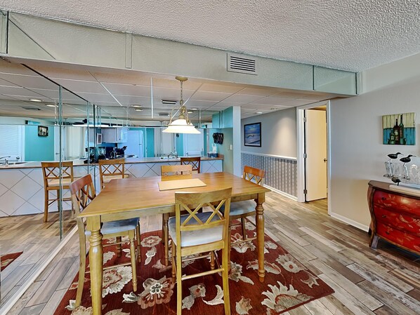 Un comedor con una mesa y sillas de madera, una alfombra y una cocina al fondo.