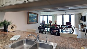 Een moderne keuken met granieten aanrechtbladen, roestvrijstalen spoelbakken en een eethoek met een glazen tafel en stoelen.