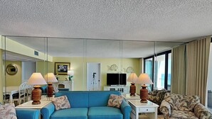 A living room with a blue sofa, a patterned armchair, and a white coffee table. There are two lamps on side tables, a television, and a tripod in the background. - Edgewater Tower II #510 | Gulf Front with Resort Amenities (Panama City Beach)
