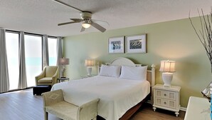 A hotel room with a large bed, a ceiling fan, two bedside lamps, a chair, a nightstand, and a view of the ocean through large windows. - Edgewater Tower II #510 | Gulf Front with Resort Amenities (Panama City Beach)