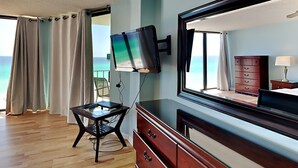 A hotel room with a large window, a flat-screen TV mounted on the wall, a wooden dresser, a small table, and a chair. - Edgewater Tower II #603 | Gulf-Front View + Amenities! (Panama City Beach)