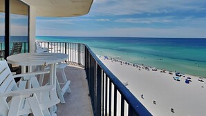 A balcony with a table and chairs, overlooking a beach with clear turquoise water and a sandy shore.