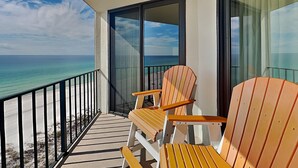 Un balcón con dos sillas Adirondack de madera, una puerta de vidrio que da al océano y vista a la playa.