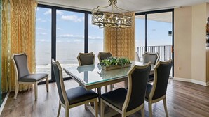 A dining area with a glass table and chairs, a chandelier, and a view of the ocean. - Edgewater Tower II #912 | Fantastic Amenities + Views! (Panama City Beach)