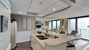Una cocina moderna con una isla central, un comedor con sofá y sillas, y un amplio ventanal con vista al mar.