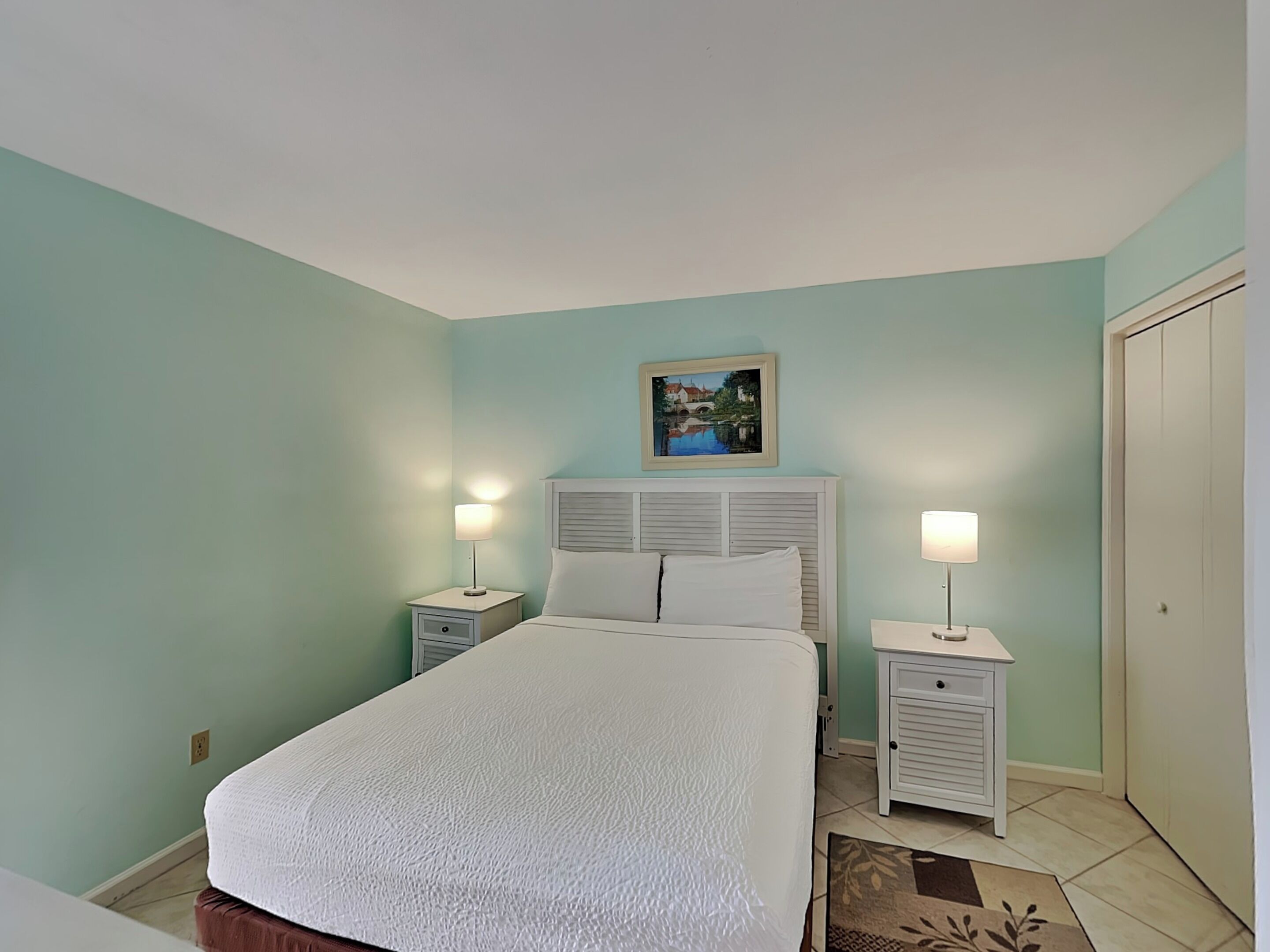 Un dormitorio con una cama, dos mesitas de noche con lámparas, una mesita auxiliar, un cuadro en la pared y una puerta.