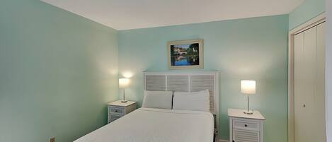 Un dormitorio con una cama, dos mesitas de noche con lámparas, una mesita auxiliar, un cuadro en la pared y una puerta.
