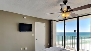 A bedroom with a bed, a ceiling fan, a TV mounted on the wall, and a door leading to a balcony with a view of the ocean. - Edgewater Tower III #1114 | Gulf Front + Wraparound Balcony (Panama City Beach)