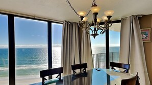 A dining area with a glass table and chairs, large windows with ocean views, and a chandelier. - Edgewater Tower III #1114 | Gulf Front + Wraparound Balcony (Panama City Beach)
