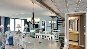 Un vaste espace de vie comprenant une table à manger, des chaises blanches et un lustre. On aperçoit un canapé et un téléviseur au fond de la pièce.
