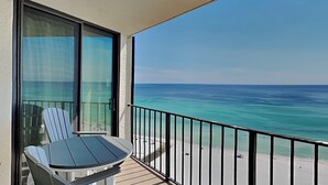 Un balcon avec une table et des chaises, donnant sur une plage où l’eau est d’un bleu turquoise cristallin et le rivage est sablonneux.